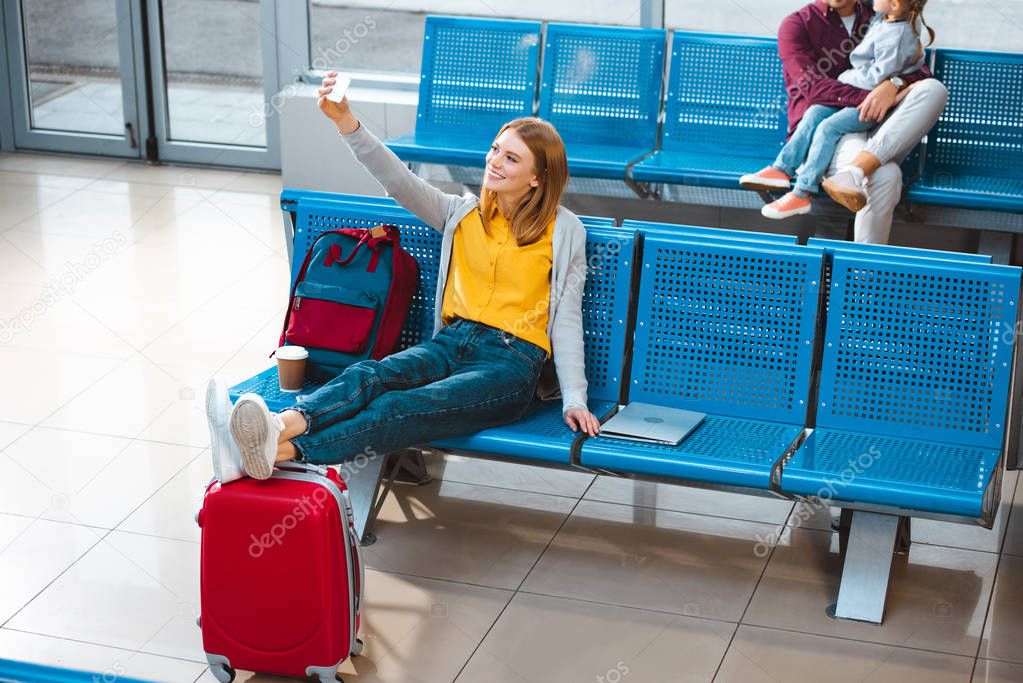 selective focus of smiling woman taking selfie in airport with people on background 