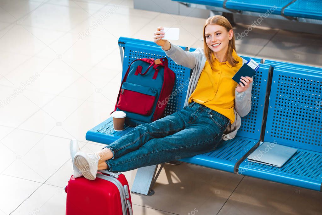 smiling woman taking selfie in airport near backpack and luggage