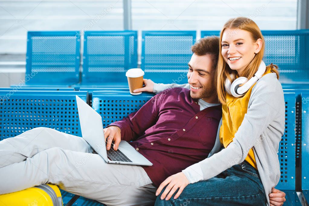 cheerful woman holding paper cup near boyfriend with laptop while sitting in airport