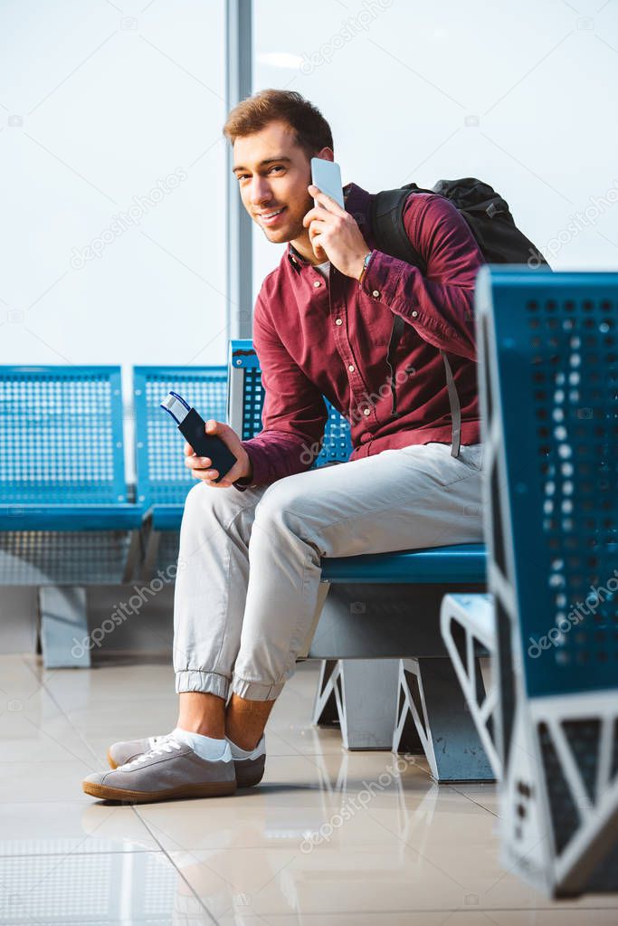 cheerful man talking on smartphone and smiling while sitting in waiting hall
