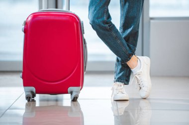 cropped view of woman standing with suitcase with crossed legs in airport  clipart