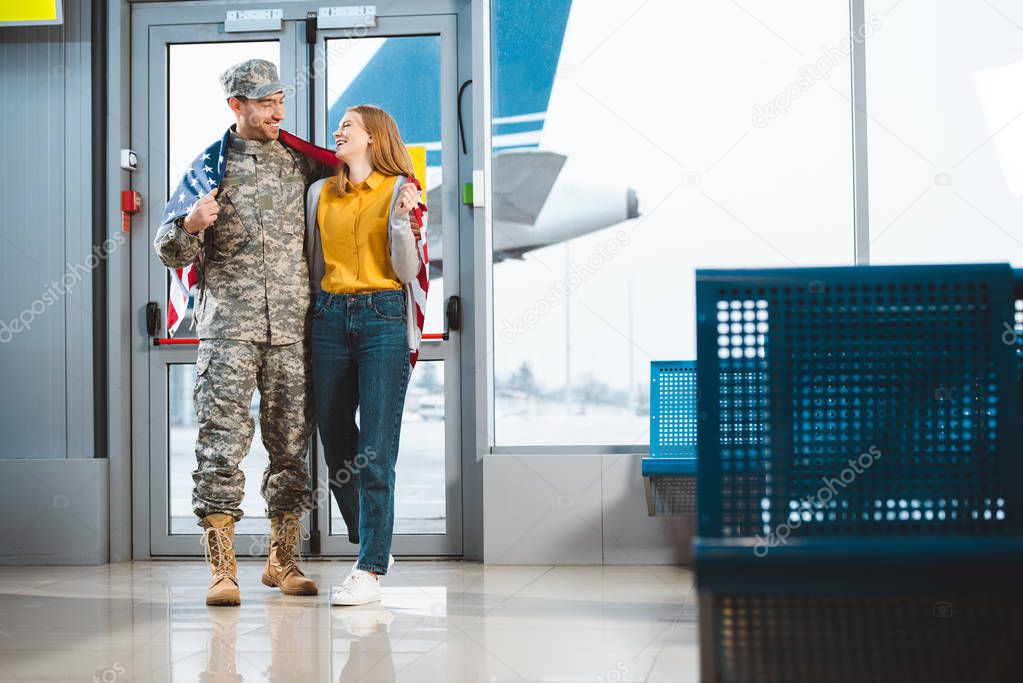 cheerful veteran in military uniform standing with girlfriend and holding american flag in airport 