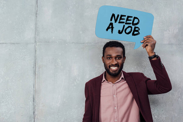 smiling african american casual businessman looking at camera and holding speech bubble with need a job lettering 