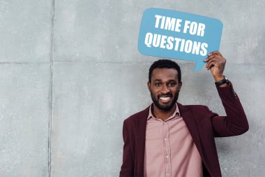 smiling african american casual businessman looking at camera and holding speech bubble with time for questions lettering clipart