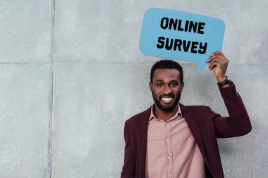 smiling african american casual businessman looking at camera and holding speech bubble with online survey lettering clipart