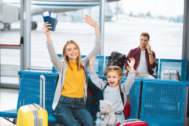selective focus cherful mother and daughter holding hands above head and smiling in airport with man on background clipart