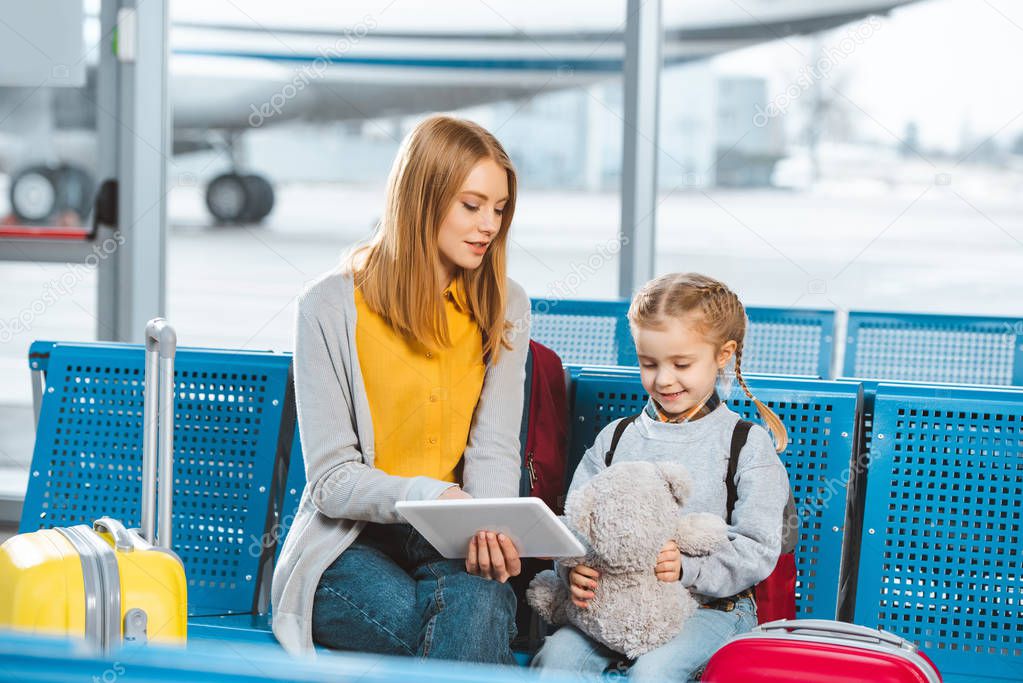 mother holding digital tablet and sitting in waiting hall with daughter 