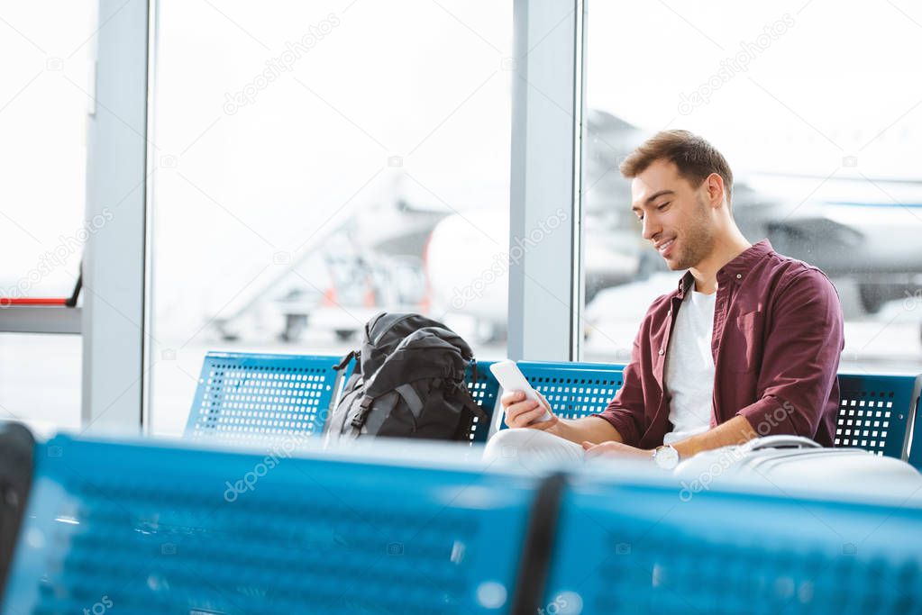 cheerful man holding smartphone while sitting in departure lounge