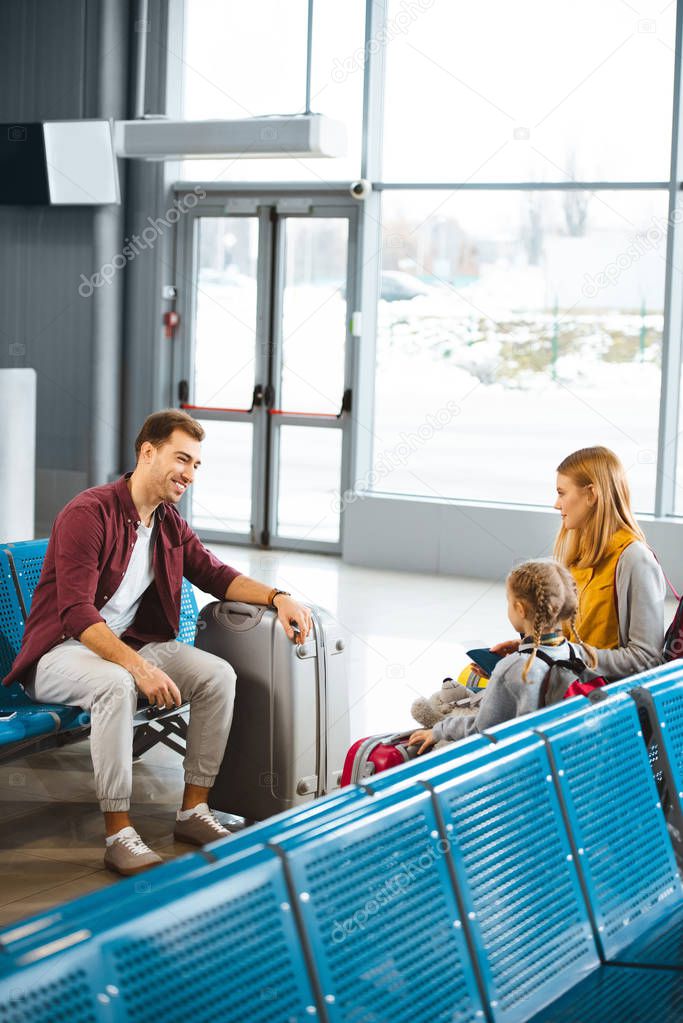 mother sitting with daughter and looking at smiling husband in airport  