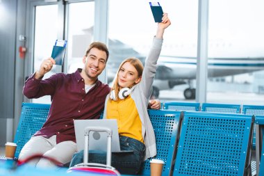 happy girlfriend holding passport above head near smiling boyfriend in airport near luggage  clipart