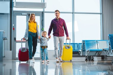 cheerful husband and wife holding hands with cute daughter and standing with suitcases clipart
