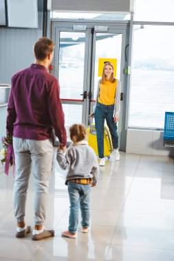 back view of man holding hands with daughter and looking at wife with luggage  clipart