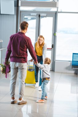 mother with baggage looking at husband and daughter holding hands while meeting in airport  clipart