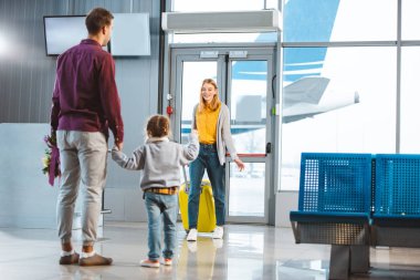 selective focus of mother with baggage looking at husband and daughter holding hands while meeting in airport  clipart