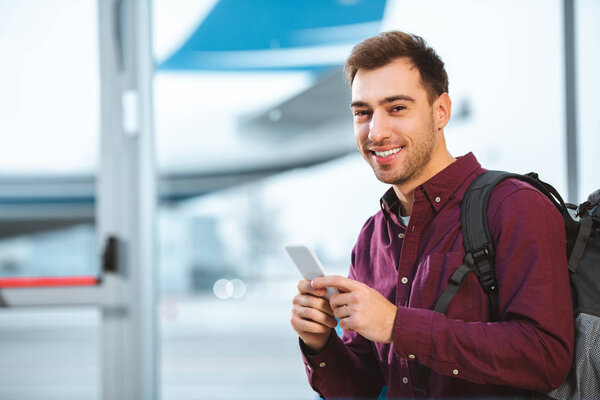 cheerful man holding smartphone and smiling in airport 