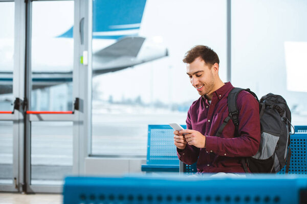cheerful man using smartphone and smiling while waiting in departure lounge