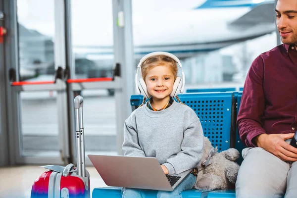 微笑的孩子听音乐在耳机 而使用笔记本电脑附近的爸爸在机场 — 图库照片