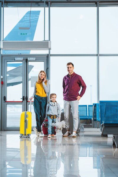 adorable child holding teddy bear and standing between mother and father in departure lounge