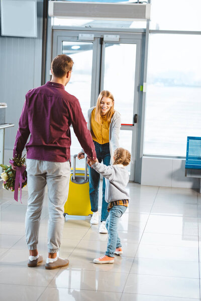 mother with baggage looking at husband and daughter holding hands while meeting in airport 