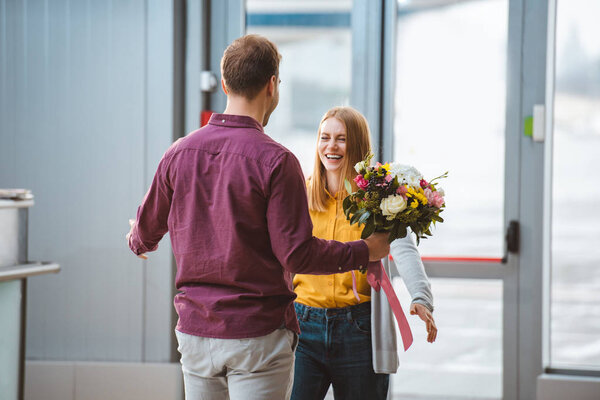 back view of man meeting cheerful girlfriend with bouquet in airport 