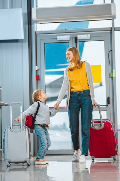 cheerful mother and daughter looking at each other while holding hands in airport 