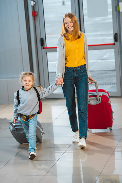 attractive mother holding hands with cute daughter and walking with baggage in airport 