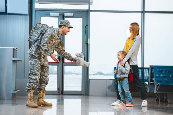 dad in military uniform giving teddy bear to daughter standing near mother in airport 