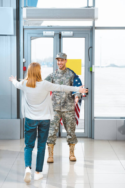 back view of woman meeting boyfriend in military uniform with american flag in airport 
