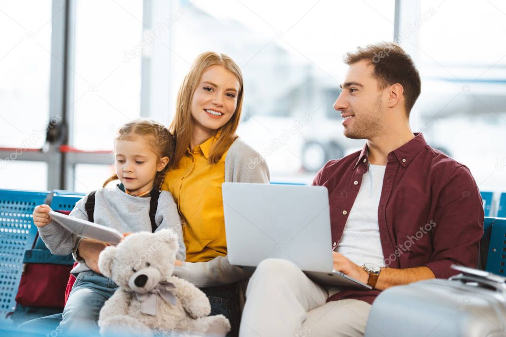 cute daughter holding digital tablet near mother and sitting near father with laptop in airport 