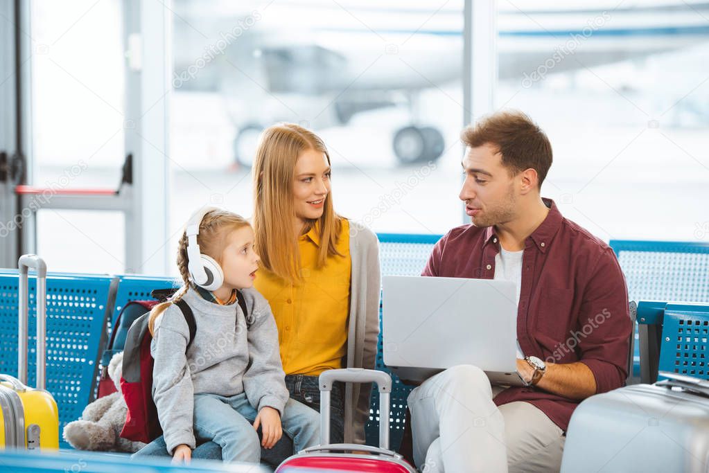 surprised kid in headphones looking at dad while sitting near mother in airport 