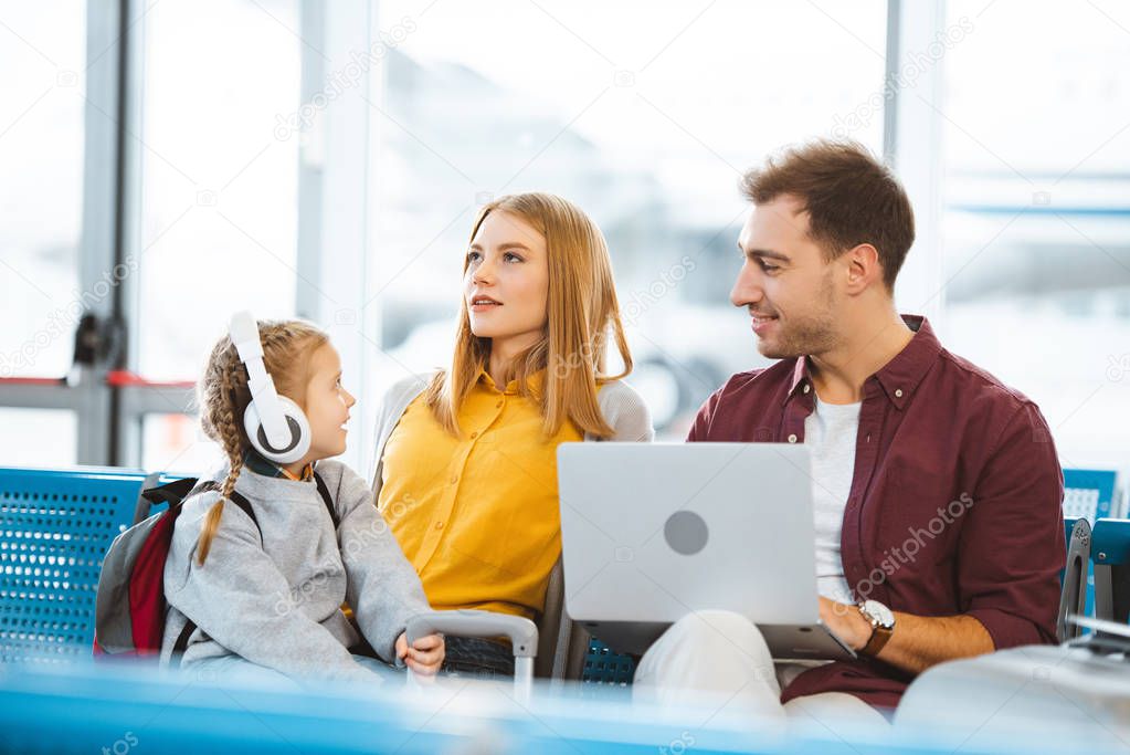 cute kid in headphones looking at mother while sitting near father in airport 