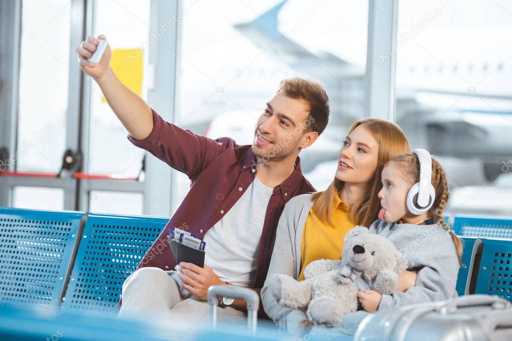 cheerful dad taking selfie and smiling near wife and daughter showing tongue in airport 