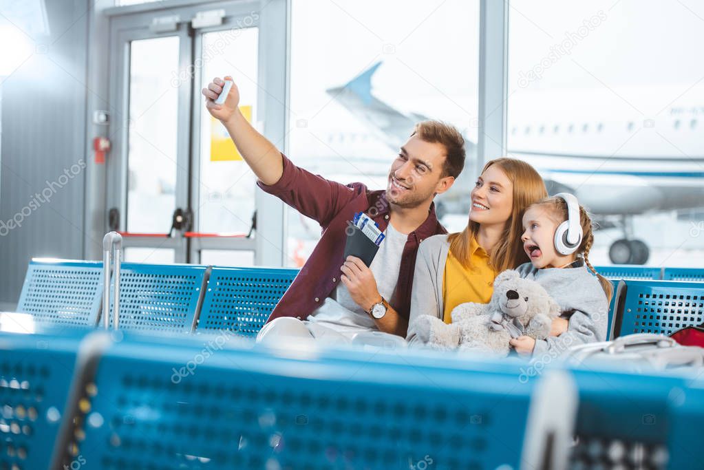 cheerful dad taking selfie and smiling with wife and daughter showing tongue in airport 