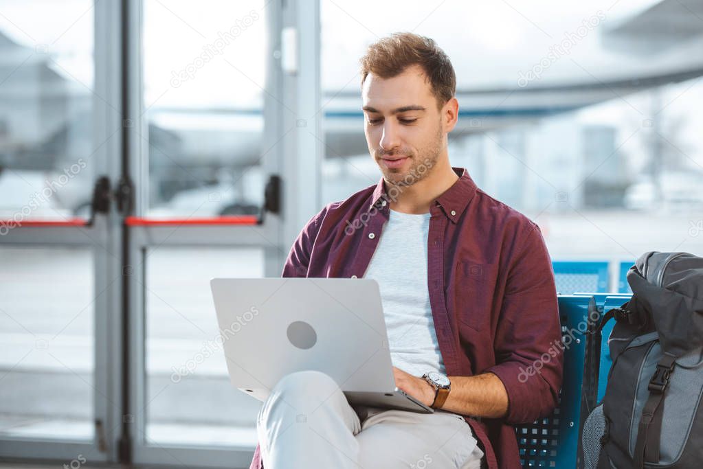handsome man looking at laptop while sitting in waiting hall
