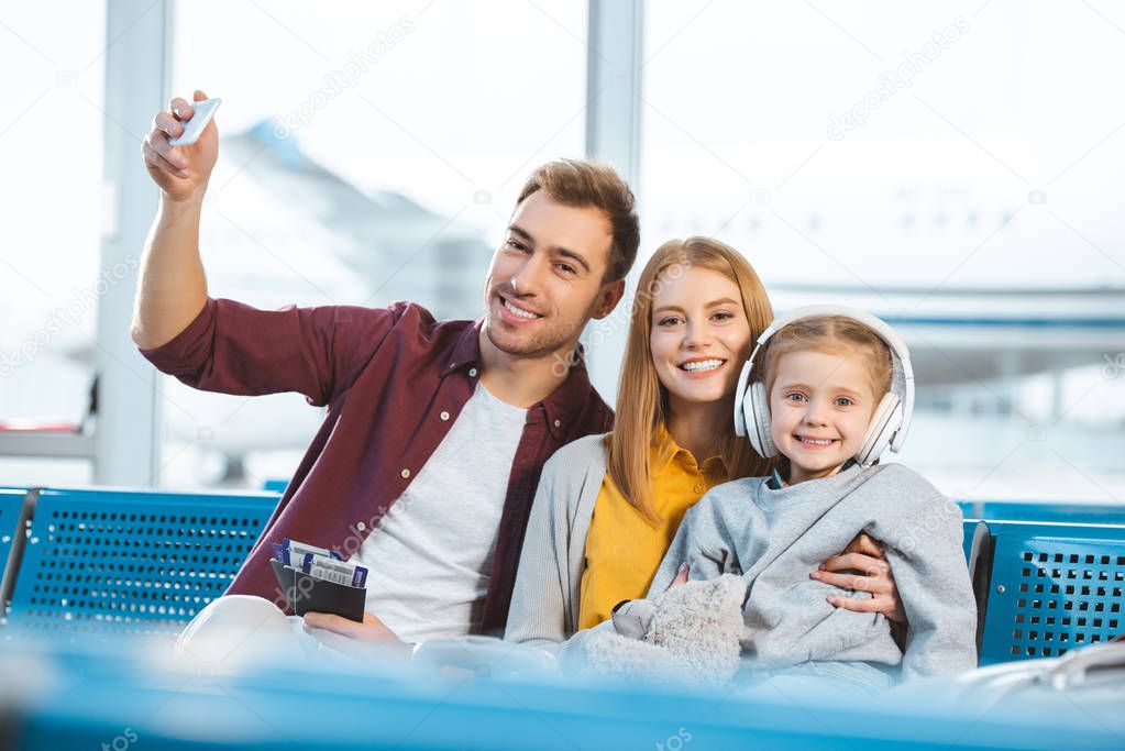 cheerful dad holding passports with air tickets and taking selfie with smiling wife and daughter 