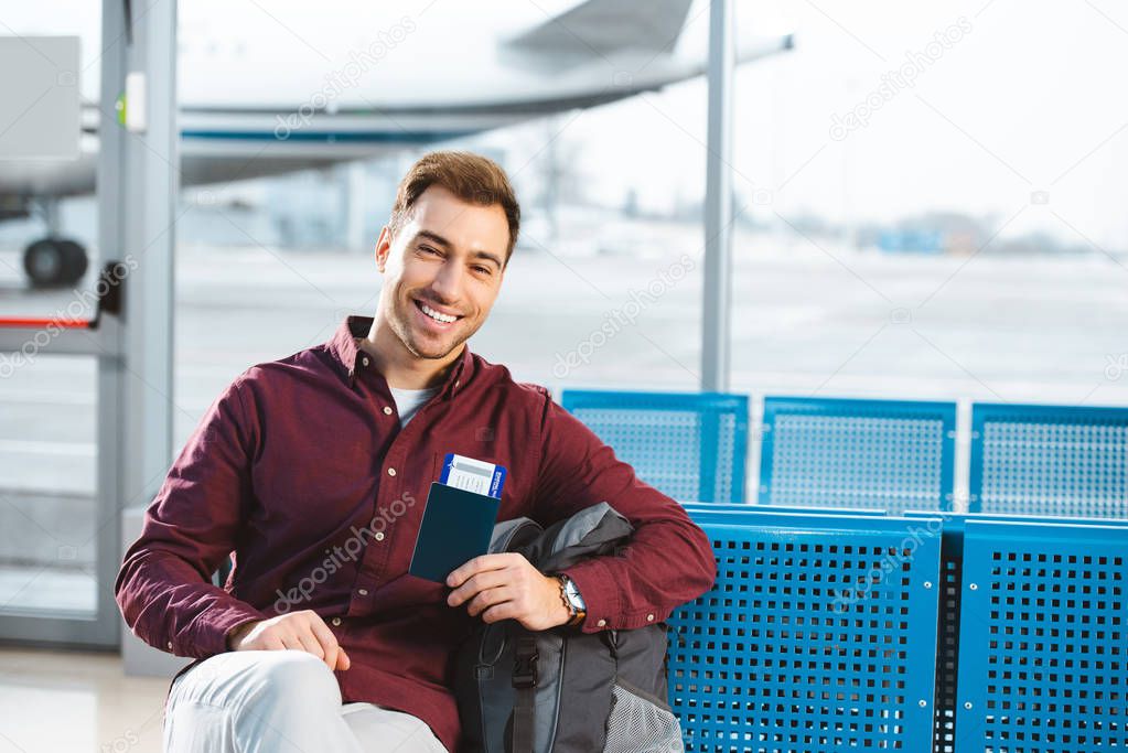 smiling man holding passport with air ticket near backpack in waiting hall