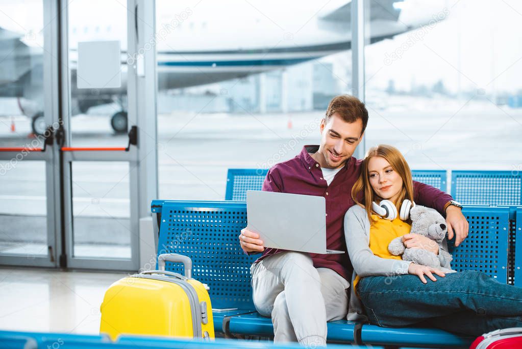 happy boyfriend holding laptop near girlfriend with teddy bear in departure lounge