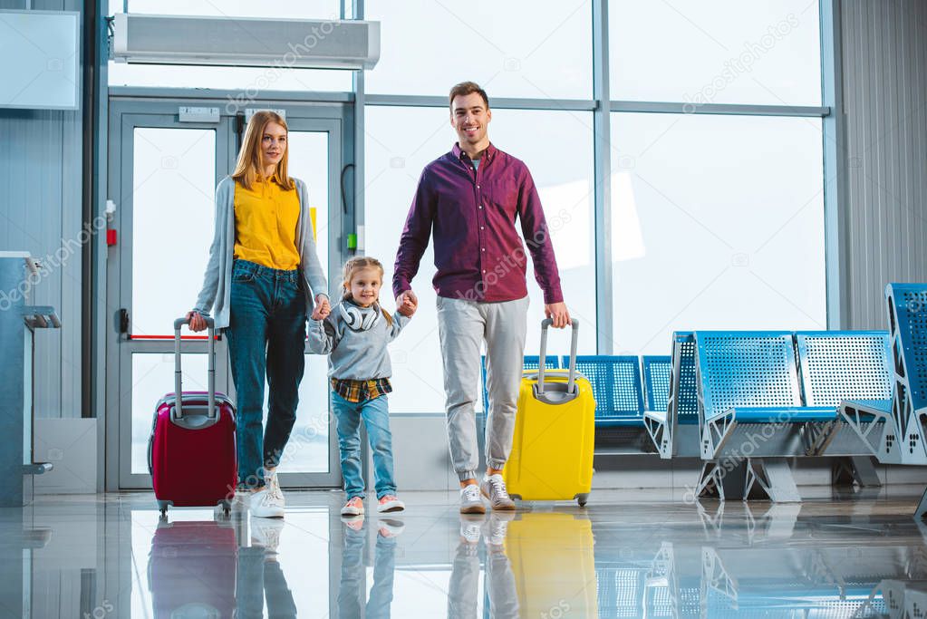 cheerful husband and wife holding hands with cute daughter and standing with suitcases