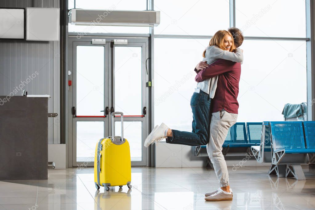 attractive woman hugging boyfriend in waiting hall near suitcase