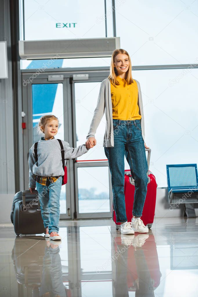 attractive mother holding hands with cute daughter while walking with suitcases in airport 