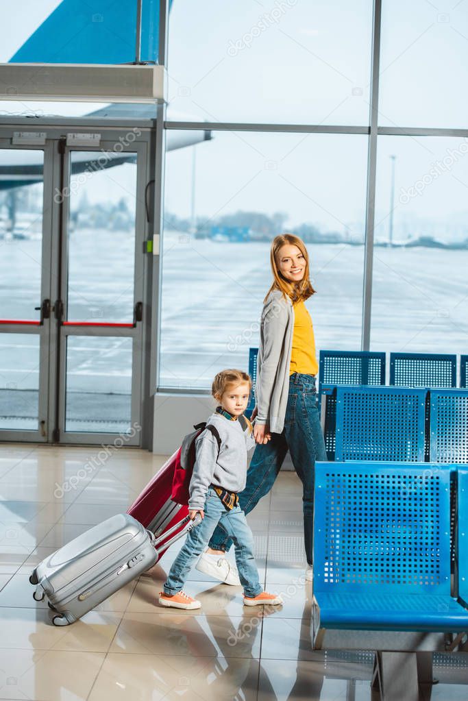 attractive mother holding hands with daughter and walking with baggage in airport 