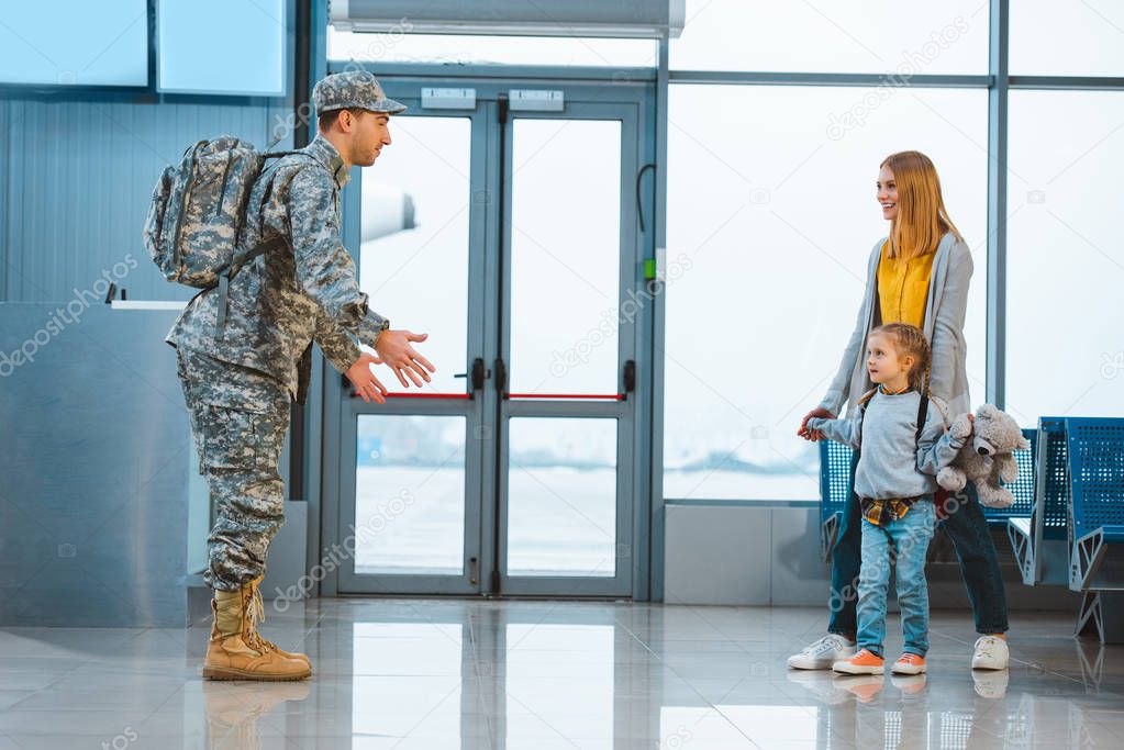 dad in military uniform standing with opened arms near wife and daughter in airport 
