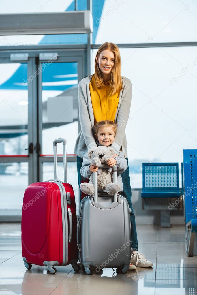 cheerful mother hugging cute daughter holding teddy bear near baggage in airport 