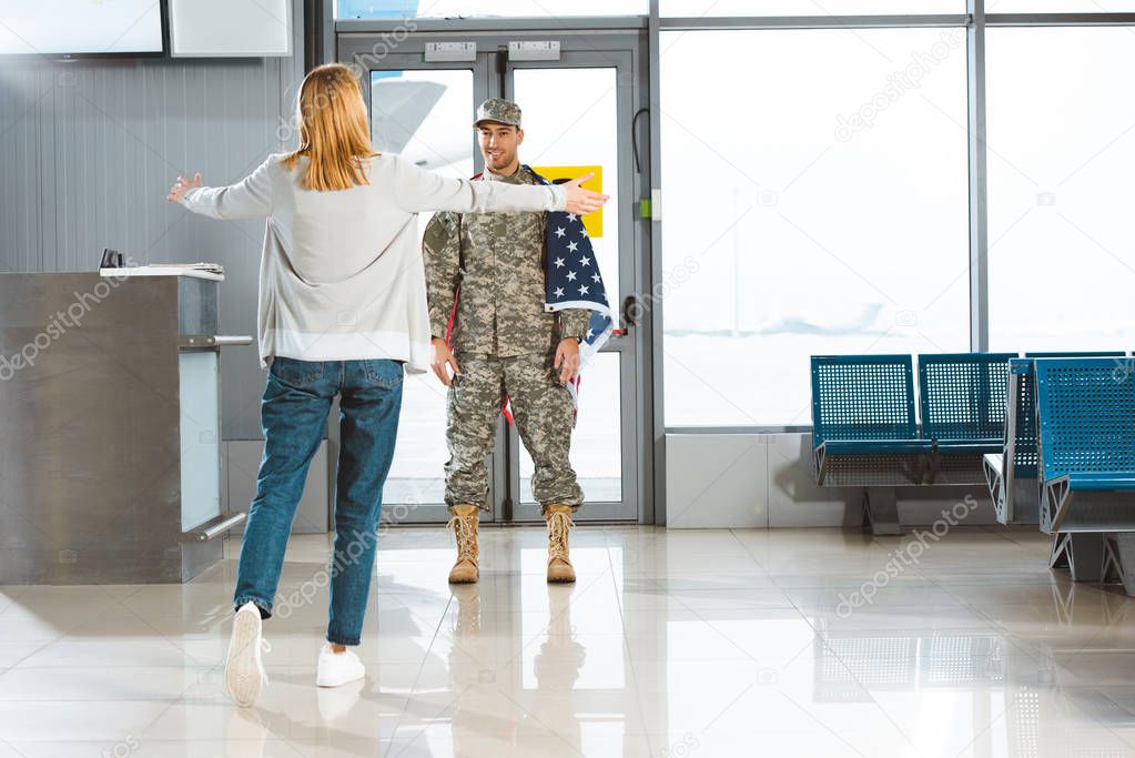 happy girlfriend with opened arms meeting boyfriend in military uniform with american flag in airport 