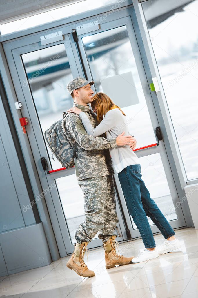 happy girlfriend hugging smiling boyfriend in military uniform in airport 