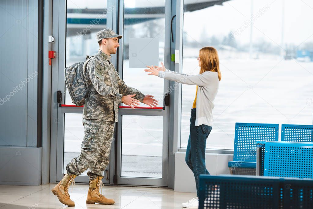happy veteran in military uniform looking at girlfriend and standing with opened arms in airport 