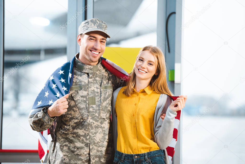 smiling man in military uniform standing with girlfriend and holding american flag in airport 