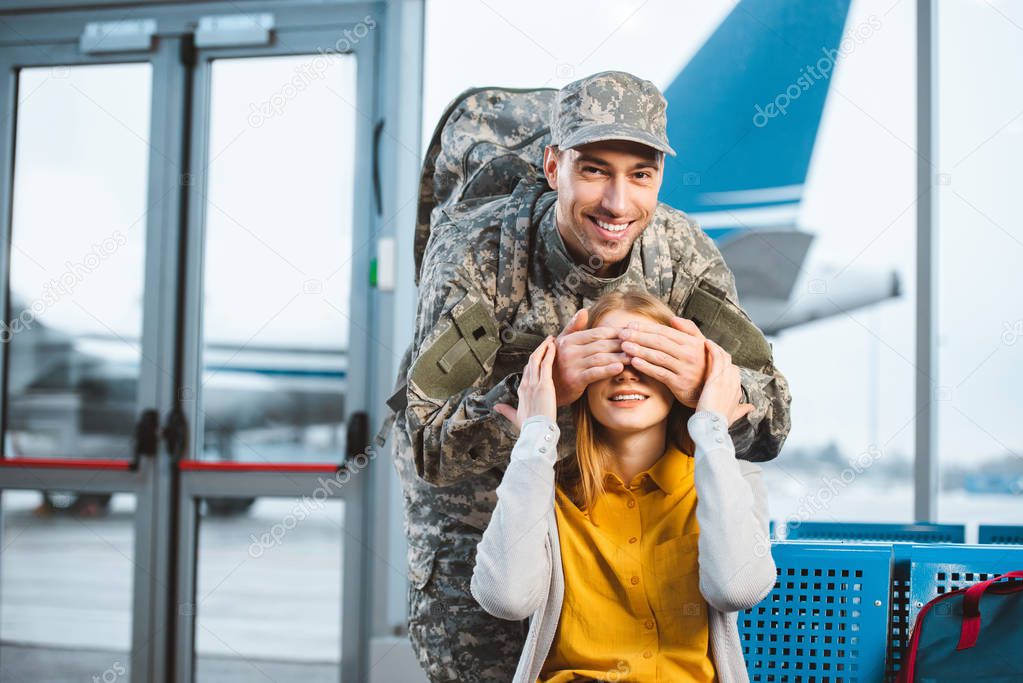handsome veteran closing eyes of cheerful girlfriend in airport 