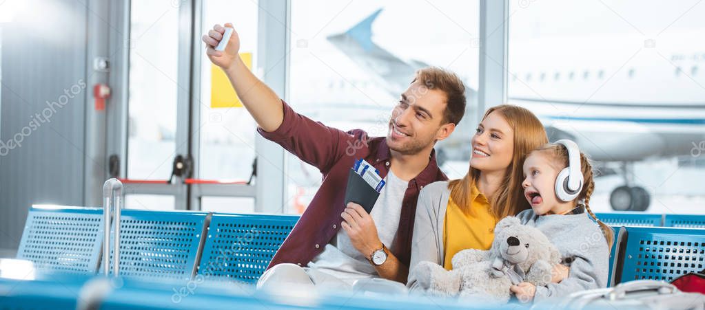 cheerful dad taking selfie and smiling with wife and daughter showing tongue in airport 