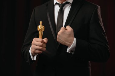 KYIV, UKRAINE - JANUARY 10, 2019: partial view of man in suit with clenched fist holding oscar award on dark background clipart